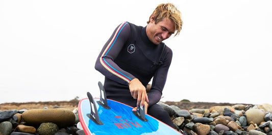 A man wearing a wet suit attaches fins to his surf board at the beach. 