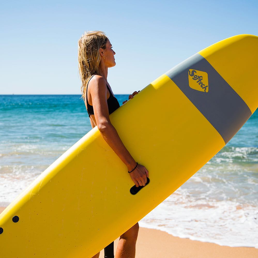 A woman holds a yellow and grey surf board on the beach. 