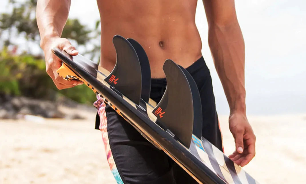 A man holds a surfboard, showing multiple black fins. 